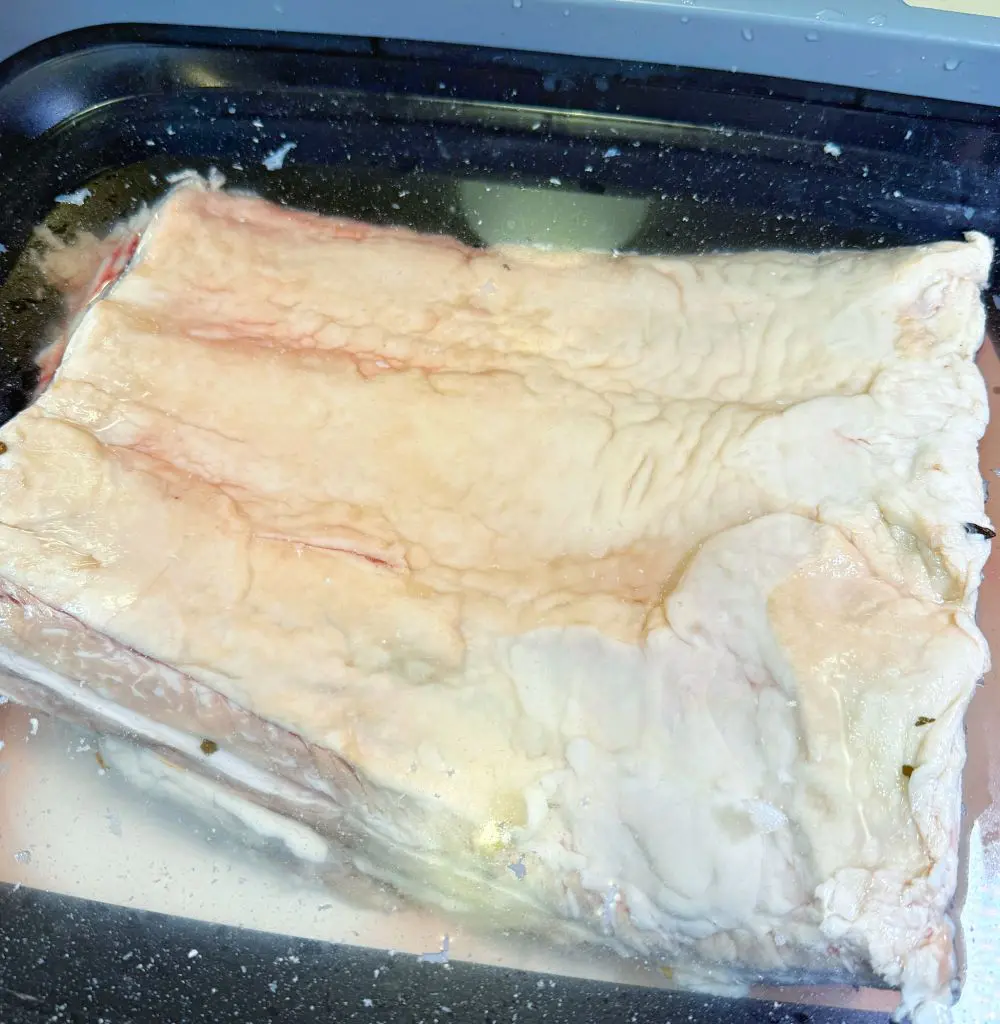 A large, raw slab of pork belly with visible fat is soaking in a tray of salted water, likely for brining or curing. The surface is pale with some red veins and wrinkles.