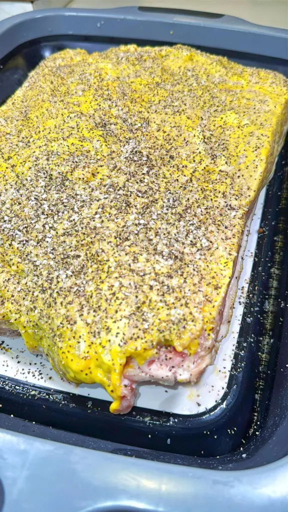 A large rectangular cut of raw meat coated with yellow mustard and heavily sprinkled with cracked black pepper sits on a tray, ready for cooking.