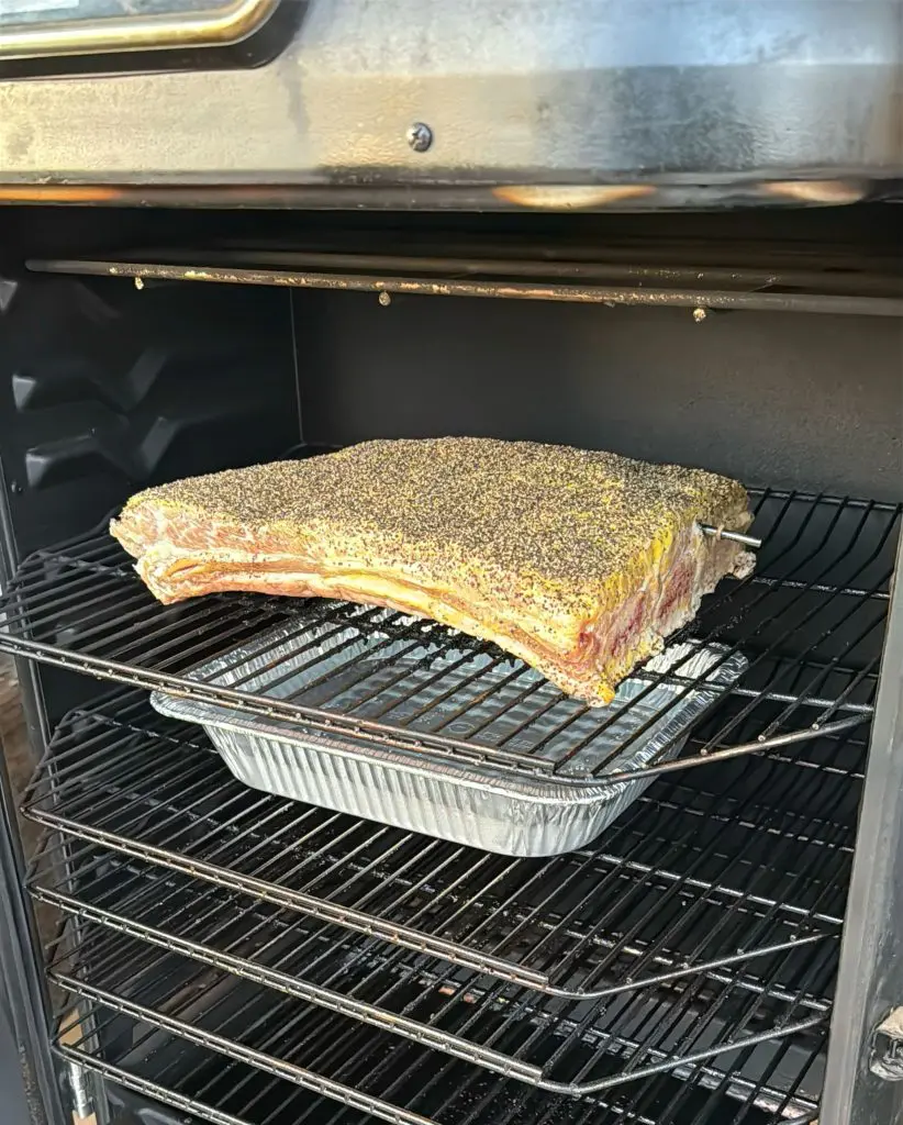 A large, seasoned brisket sits on a rack inside a smoker above a foil drip pan, ready to be cooked. The brisket is coated with a spice rub reminiscent of pastrami, and a temperature probe is inserted.