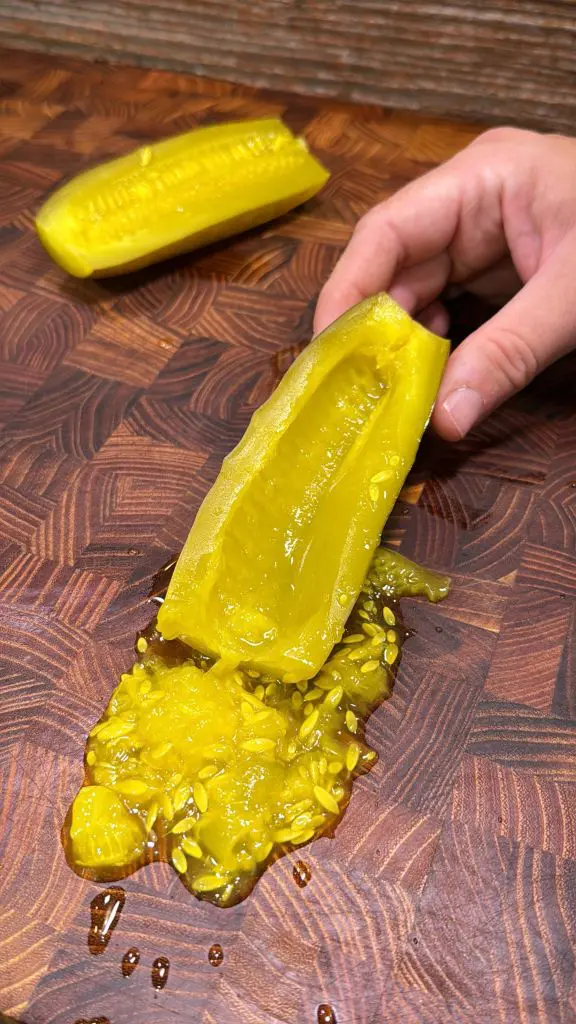 A hand holding a sliced yellow pickle on a brown wooden cutting board, with pickle seeds and juice spilling out onto the board. Another pickle half is in the background.
