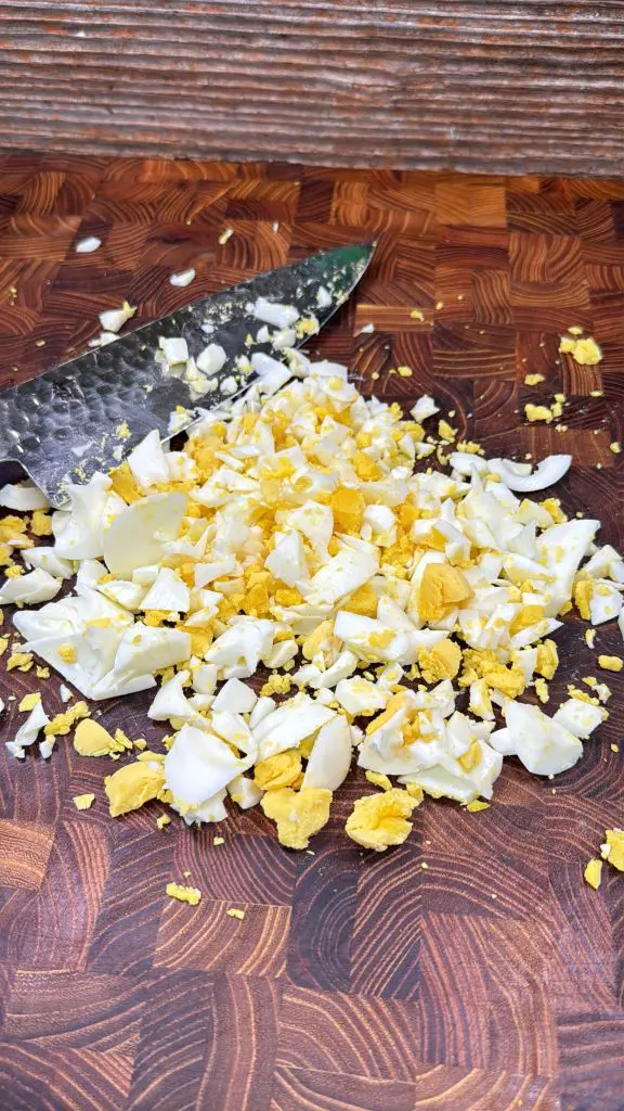 Chopped hard-boiled eggs with yellow yolks and white pieces are spread on a wooden cutting board, with a knife visible beside the eggs.