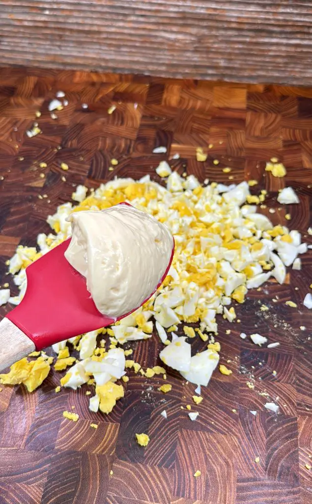 A red spatula holding mayonnaise over a pile of chopped hard-boiled eggs on a wooden cutting board.