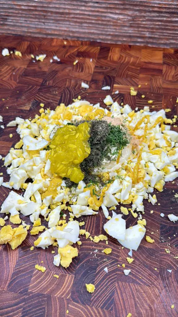 Chopped hard-boiled eggs on a wooden cutting board topped with mustard, black pepper, dried dill, and a dollop of mayonnaise, ready to be mixed for egg salad.