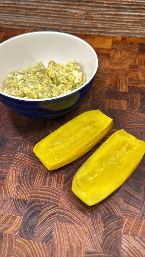 A bowl of chopped egg salad sits on a wooden cutting board next to a yellow pickle split lengthwise and hollowed out, ready for stuffing.