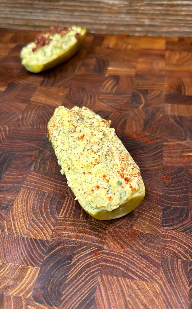 A halved pickle on a wooden board, filled with a creamy, yellowish deviled egg pickle mixture and sprinkled with paprika. Another similar filled pickle is blurred in the background.