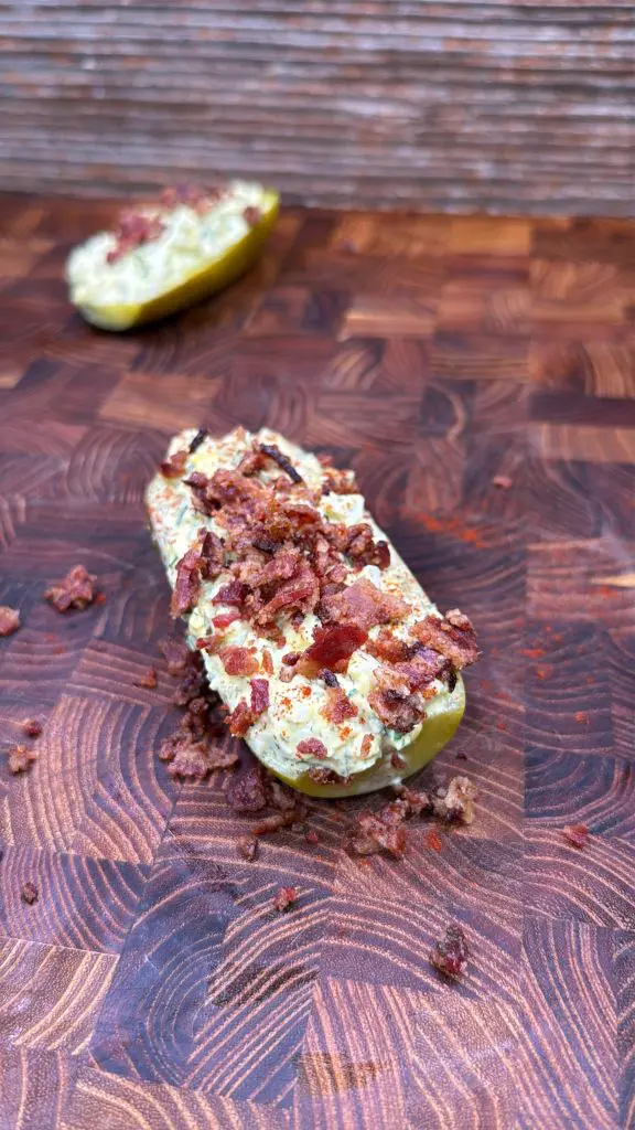A halved pickle stuffed with creamy filling and topped with crumbled bacon sits on a wooden cutting board, with another stuffed pickle half blurred in the background.