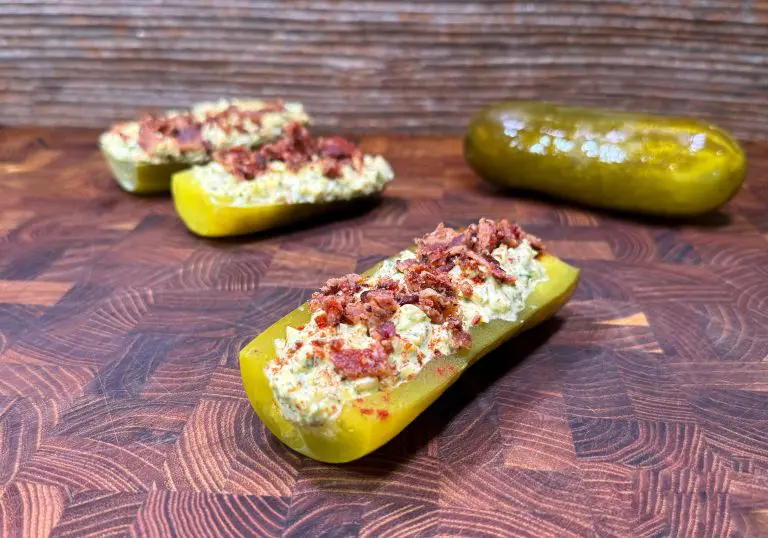 A halved pickle stuffed with a creamy filling and topped with bacon bits, placed on a wooden surface. More stuffed pickles and a whole pickle are in the background.