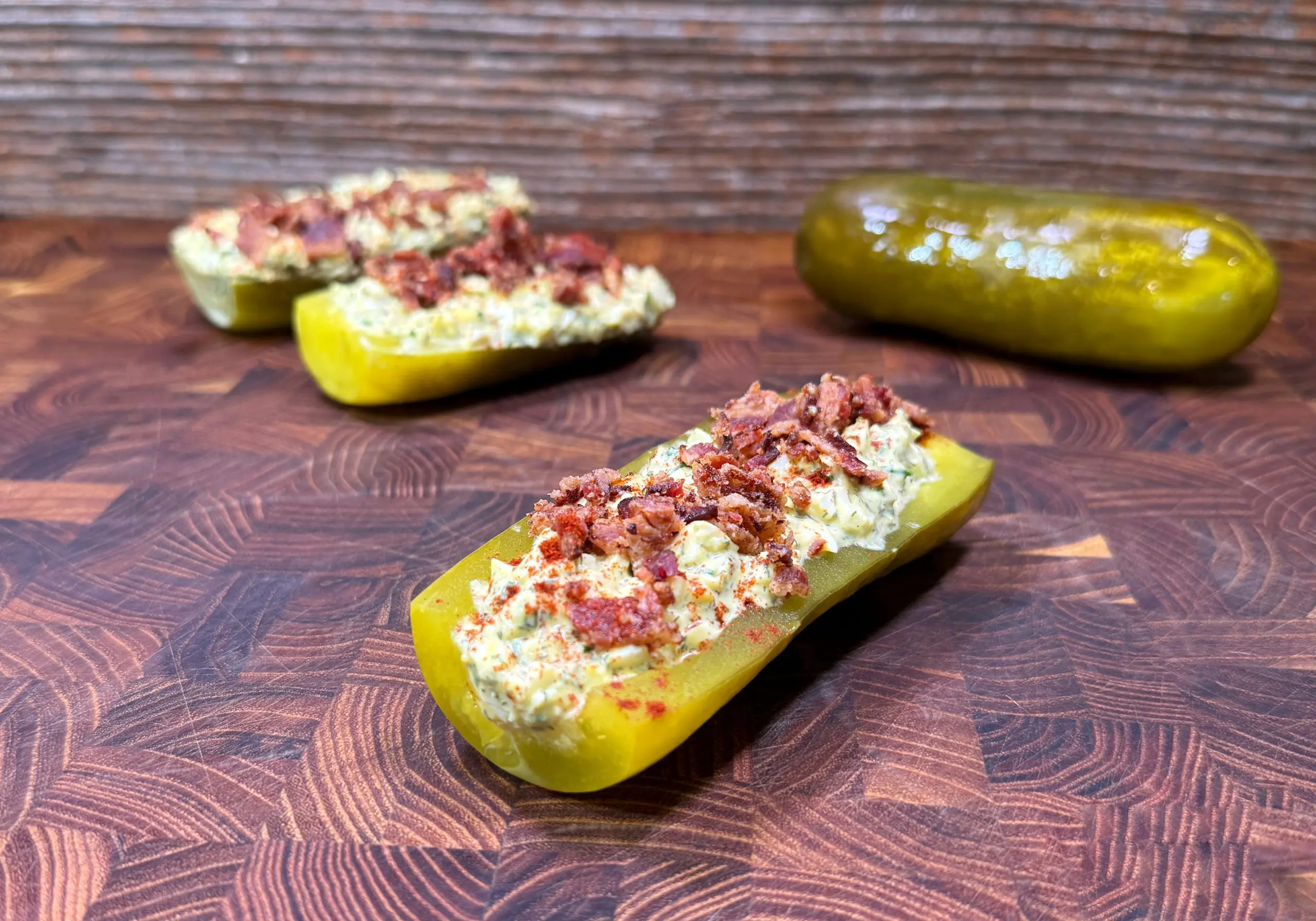 A halved pickle stuffed with a creamy filling and topped with bacon bits, placed on a wooden surface. More stuffed pickles and a whole pickle are in the background.