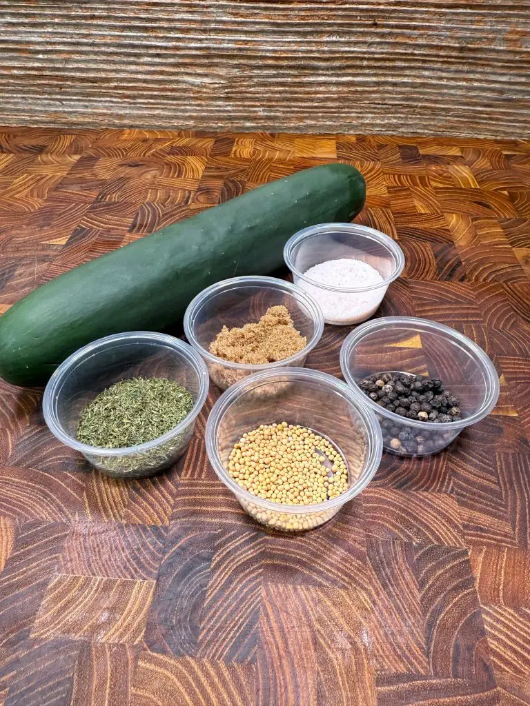A cucumber and six small plastic containers with dried dill, mustard seeds, peppercorns, brown sugar, white salt, and light brown sugar on a patterned wooden surface.