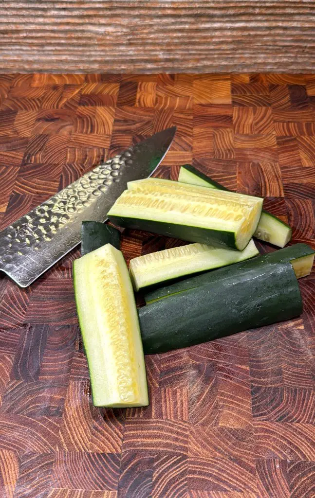 A chef’s knife and several quartered cucumber pieces are placed on a wooden cutting board with a patterned surface.