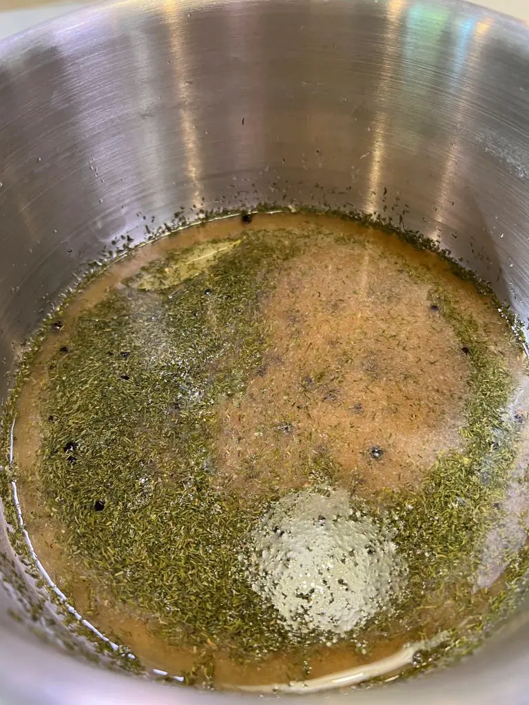 A close-up of a stainless steel pot containing a mixture of oil, herbs, and spices suspended in liquid, beer brine for the beer brine pickles, with visible flecks of seasonings floating on the surface.