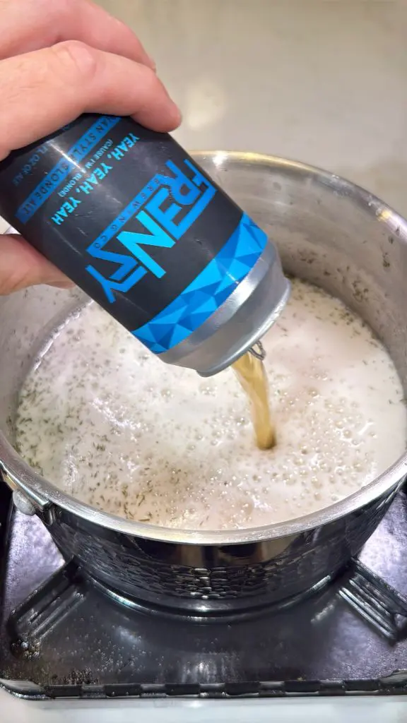 A hand pours a can of Penny IPA beer into a stainless steel pot filled with foamy, bubbling liquid on a stovetop.