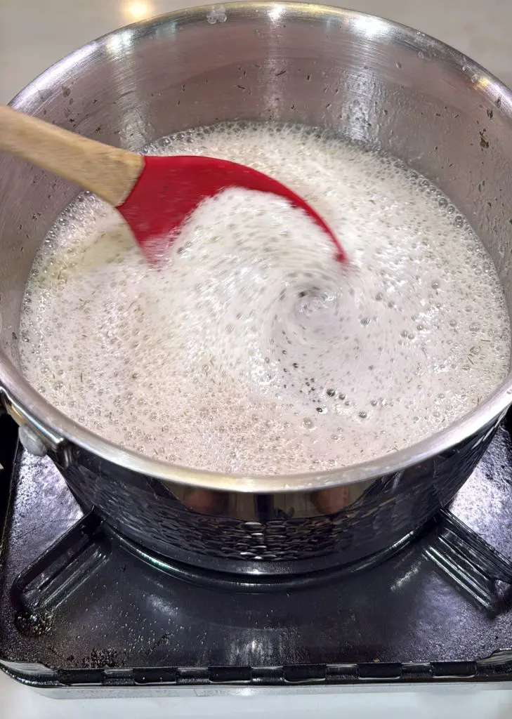 A red spatula stirs a pot of bubbling, frothy liquid on a gas stove, creating a whirlpool effect in the center.