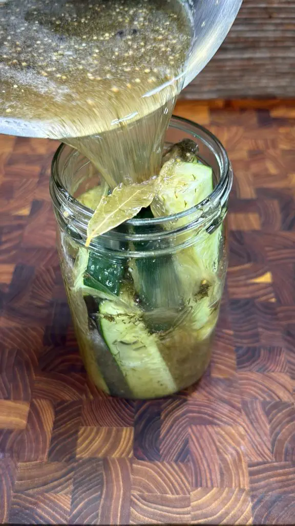 Brine being poured from a bowl into a glass jar filled with cucumber spears and bay leaves on a patterned wooden surface.