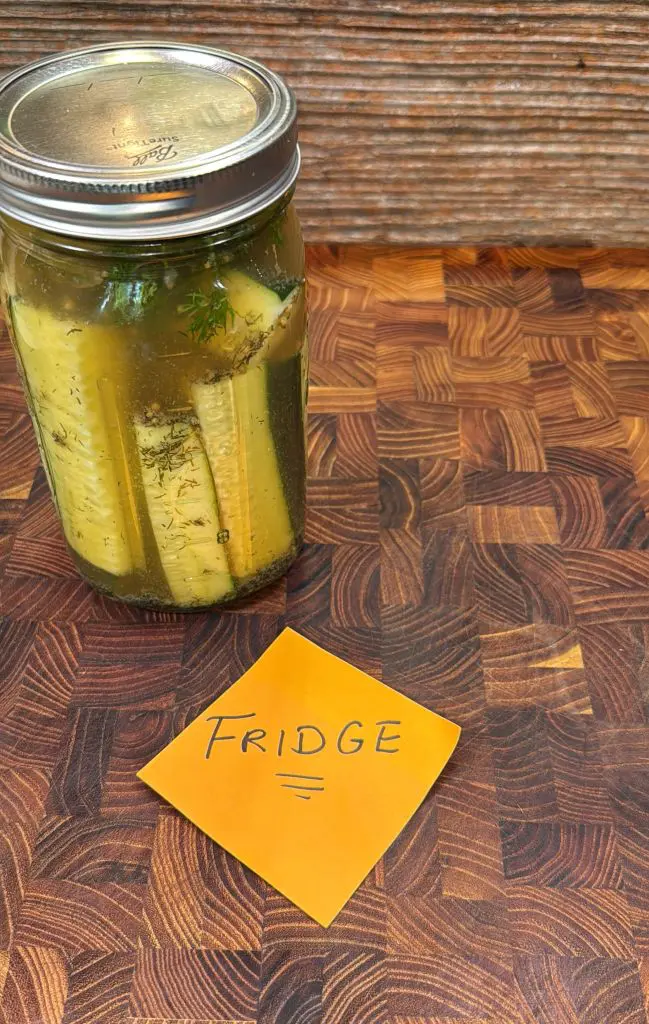 A jar of pickles sits on a wooden surface next to an orange sticky note labeled FRIDGE in large, black handwritten letters.