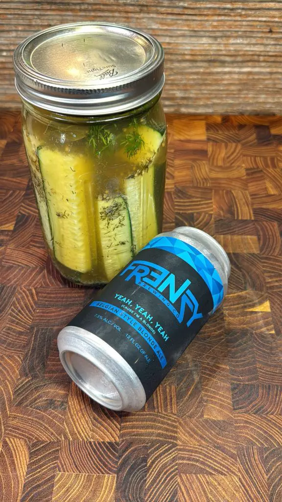 A jar of pickles with herbs inside sits next to a blue and black can labeled Frenzy on a wooden cutting board with a patterned surface.
