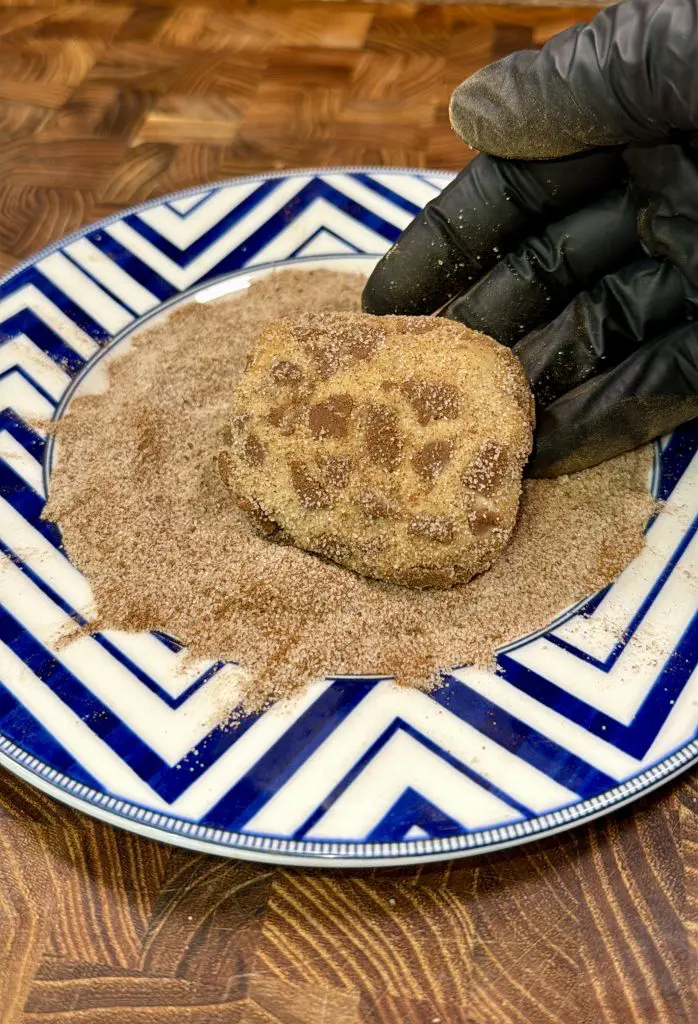 A gloved hand rolls a piece of dough in cinnamon sugar on a blue and white patterned plate, set on a wooden surface.