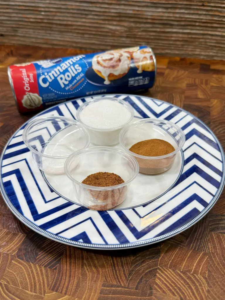 A can of cinnamon roll breakfast bombs sits behind a plate holding four small containers filled with sugar, cinnamon, nutmeg, and another spice on a chevron-patterned plate atop a wooden surface.