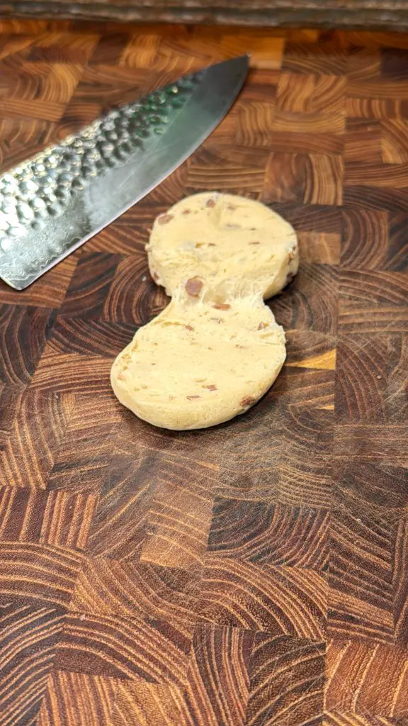 A sliced cookie dough log with visible chocolate chips rests on a wooden cutting board beside a textured metal knife, reminiscent of preparing cinnamon roll breakfast bombs for a cozy morning treat.