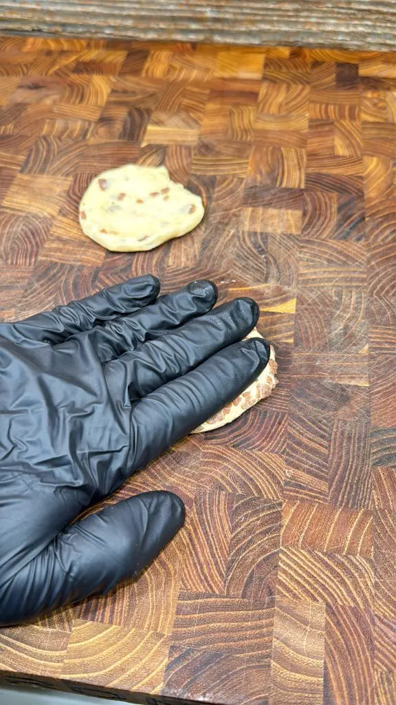 A hand wearing a black glove presses down on a small round piece of dough—soon to become cinnamon roll breakfast bombs—on a wooden cutting board, with another piece of dough in the background.