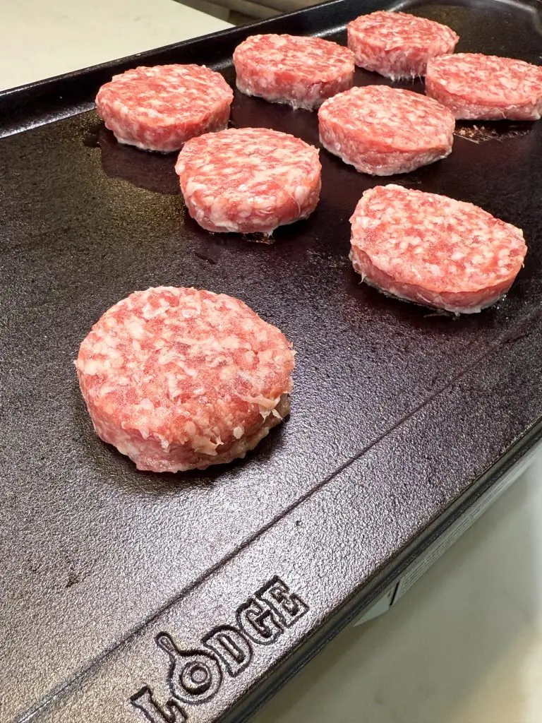 Raw sausage patties are arranged on a black Lodge griddle, ready to be cooked alongside delicious cinnamon roll breakfast bombs. The patties are evenly spaced, and the griddle surface is clean with the Lodge brand name visible in the corner.