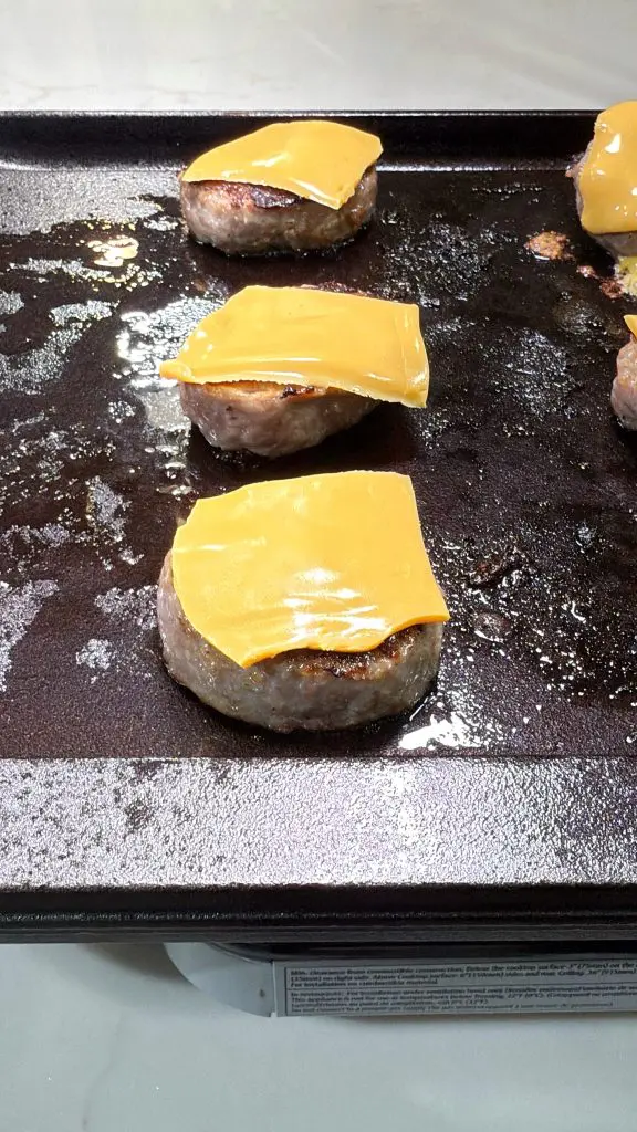 Four hamburger patties, each topped with a slice of melted cheddar cheese, are cooking on a greasy griddle. A few more patties and some cinnamon roll breakfast bombs are partly visible on the right side of the image.