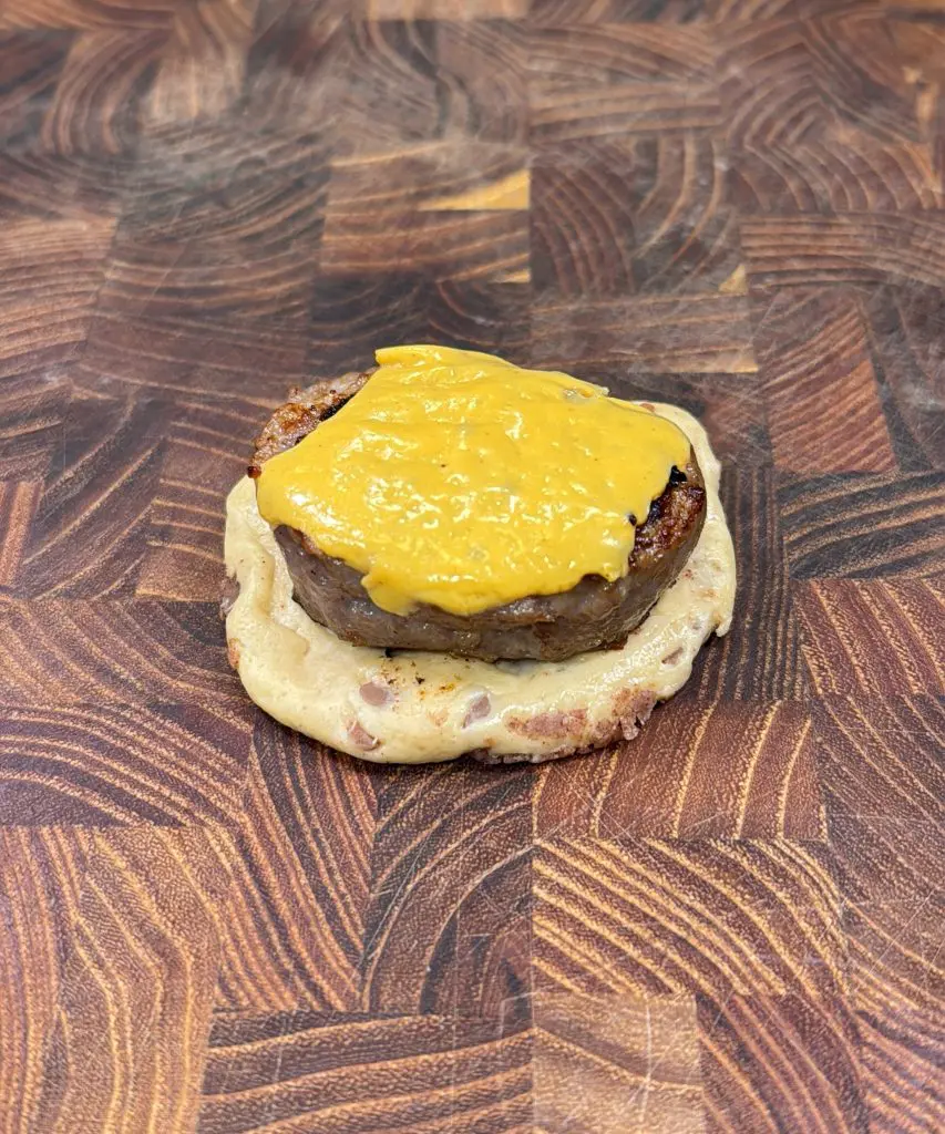 A sausage patty with melted yellow cheese sits on half of an English muffin, placed on a wooden cutting board with a patterned grain—perfect alongside cinnamon roll breakfast bombs for a delicious morning spread.