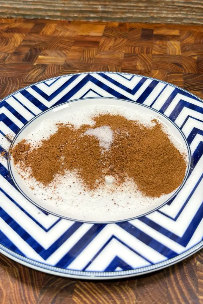 A white plate with a blue zigzag pattern holds a serving of rice pudding topped with a generous dusting of brown cinnamon, reminiscent of cinnamon roll breakfast bombs. The plate sits on a wooden surface.