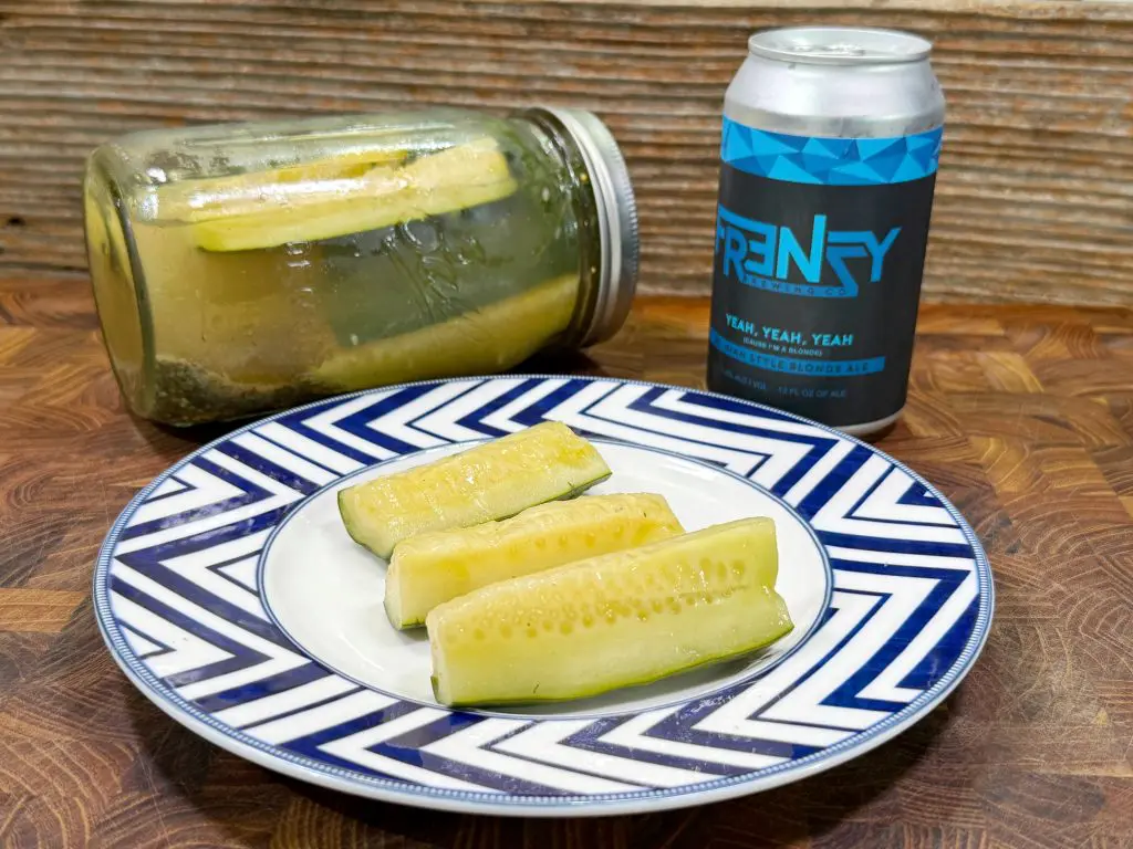 A plate with four pickle spears sits on a blue and white patterned plate. Behind it, theres a jar of pickles lying on its side and a can of Frenzy beer on a wooden surface with a rustic wood background.