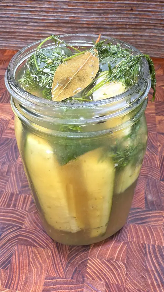A glass jar filled with pickling cucumbers, fresh dill, a bay leaf, and brine sits on a wooden surface.
