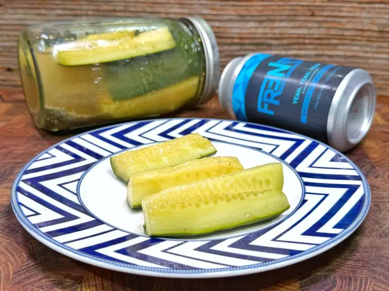 A plate with four pickle spears sits on a wooden surface. Behind the plate, there is a glass jar with pickles and a blue FREN beverage can lying on its side, all against a wood-textured background.