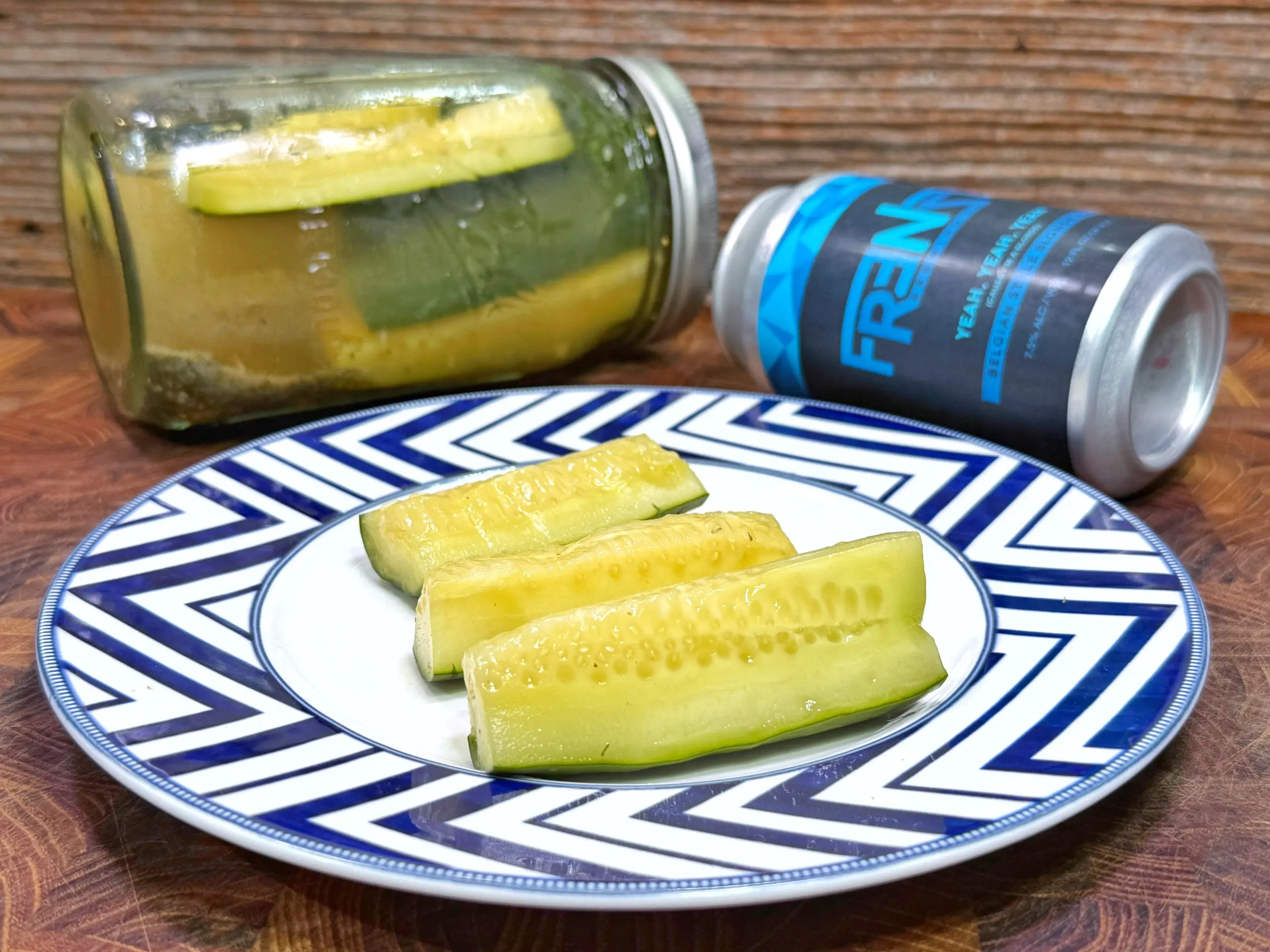 A plate with four pickle spears sits on a wooden surface. Behind the plate, there is a glass jar with pickles and a blue FREN beverage can lying on its side, all against a wood-textured background.