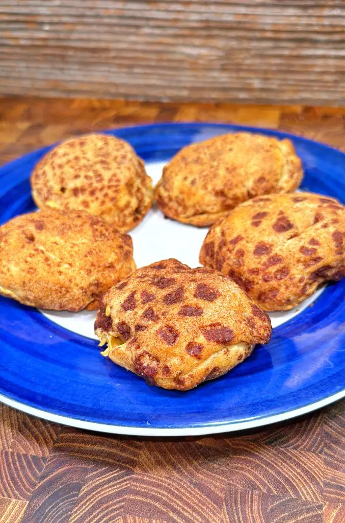 Five golden-brown baked pastry buns with a speckled appearance are arranged on a white plate with a blue rim, set on a wooden surface with a textured brown background.