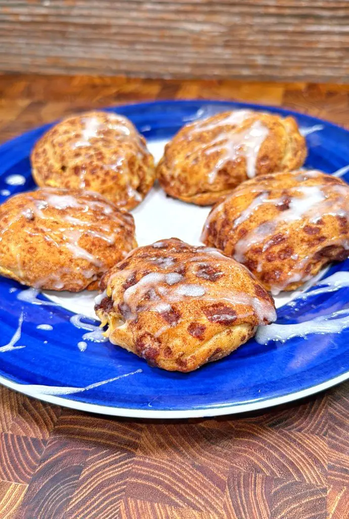 Five golden-brown cinnamon rolls with icing drizzle are arranged on a blue and white plate, set on a wooden surface.