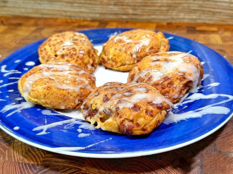 Five iced cinnamon rolls with a drizzle of white glaze are arranged on a blue and white plate, set on a wooden surface with a wood-patterned background.
