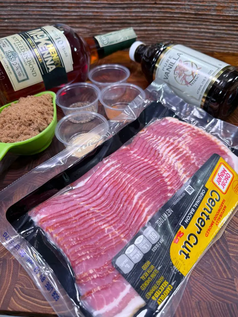 A package of center cut bacon, a cup of brown sugar, spices in small containers, a bottle of bourbon, and a bottle of vanilla extract are arranged on a wooden surface.
