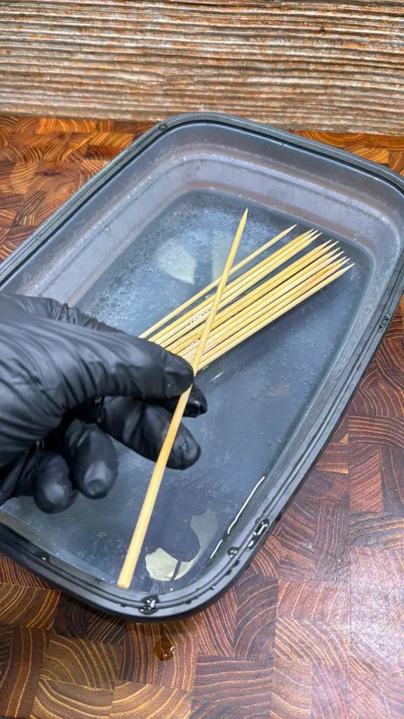 A gloved hand holds a wooden skewer above a plastic container filled with water, where several skewers are soaking on a wooden surface.