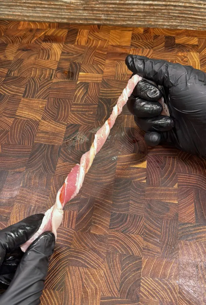 Two hands wearing black gloves twist a strip of bacon around a thin skewer on a patterned wooden cutting board.