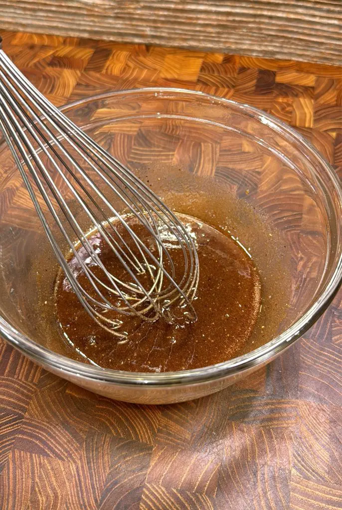A metal whisk is resting in a glass bowl filled with a brown, glossy liquid mixture on a wooden countertop with a patterned surface.