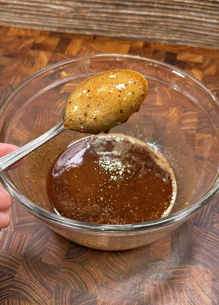 A hand holds a spoon with a thick, brown liquid mixture above a glass bowl containing more of the same mixture, on a wooden surface.