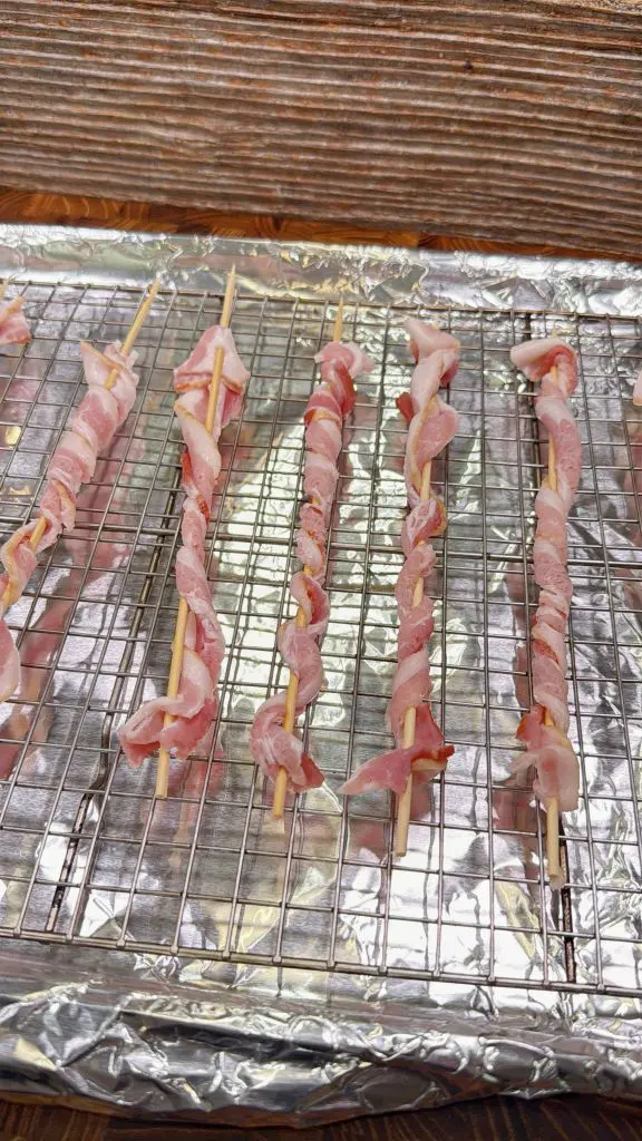 Raw bacon strips are twisted around wooden skewers and placed in rows on a wire rack over a foil-lined baking sheet, ready for cooking.
