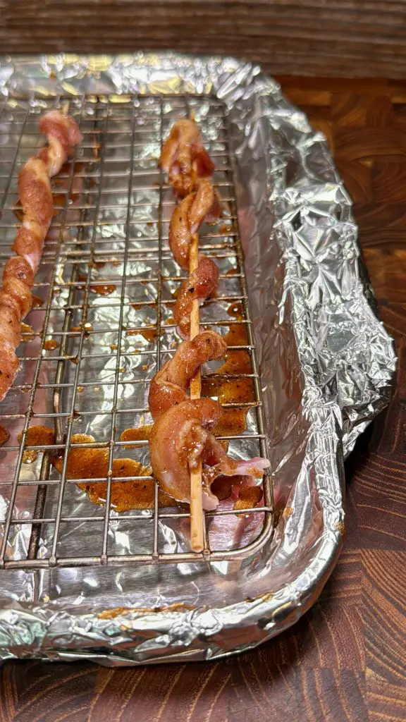 Raw bacon twisted on skewers sits on a wire rack over a foil-lined baking tray, ready to be cooked. The setup is placed on a wooden surface.