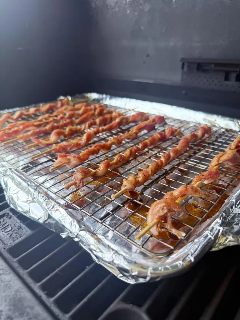 Skewers of bacon are lined up on a wire rack inside a grill or smoker. The rack sits over a foil-covered tray, with the bacon cooking and fat dripping below.