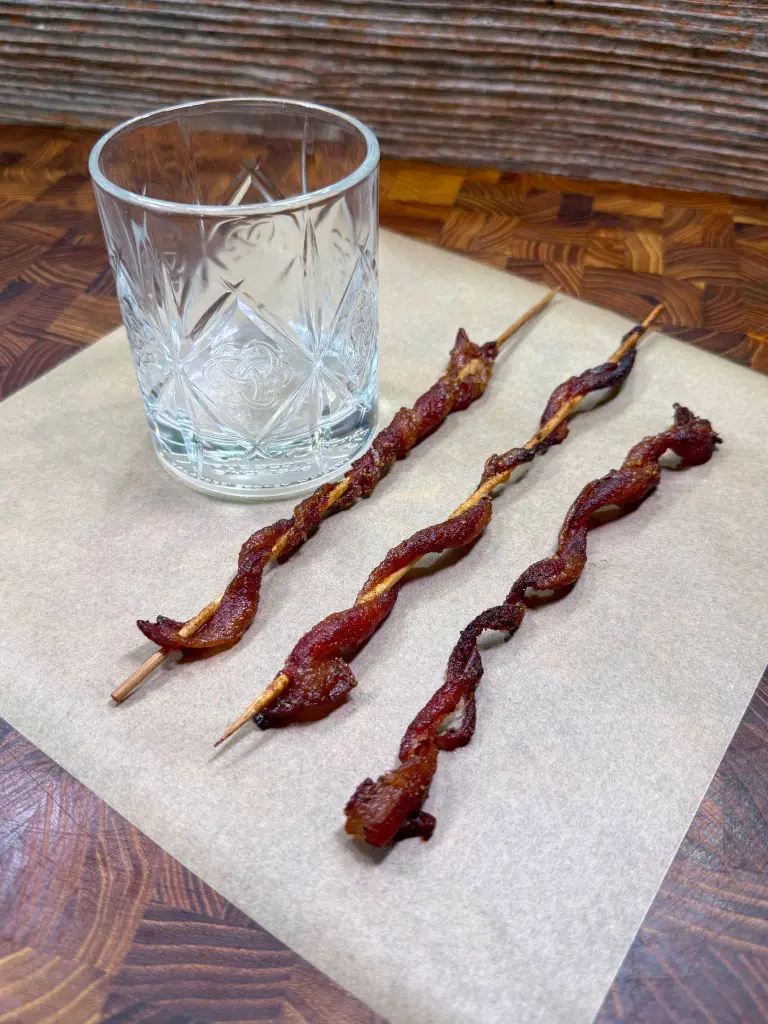A clear, empty glass tumbler sits next to three skewers of twisted, cooked bacon on parchment paper atop a wooden surface.