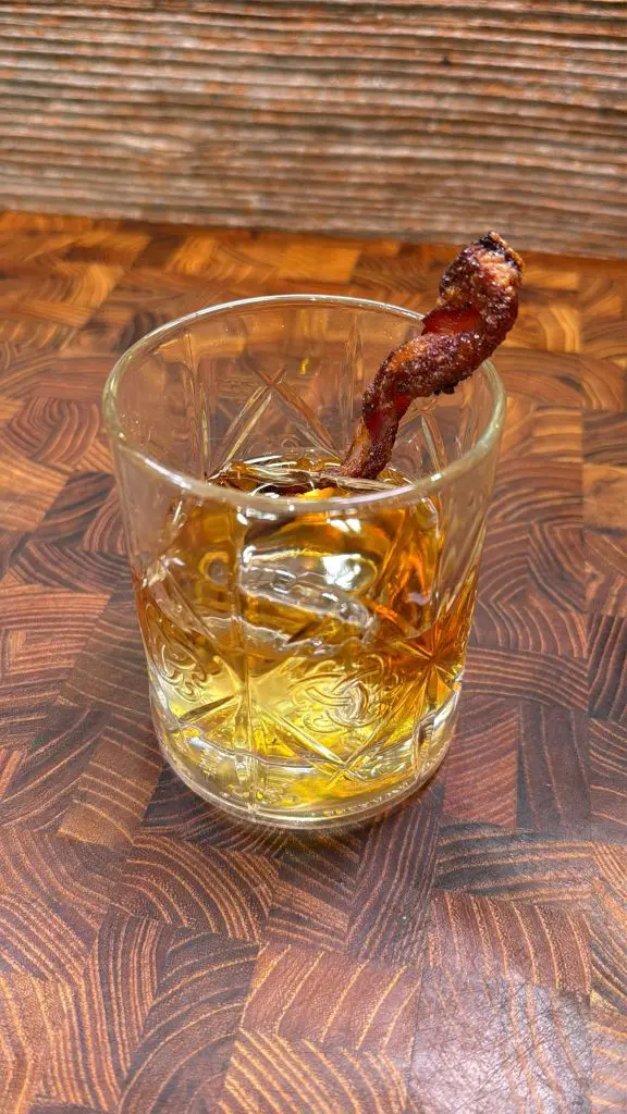 A glass of whiskey sits on a wooden surface, garnished with a crispy strip of bacon inside the drink.