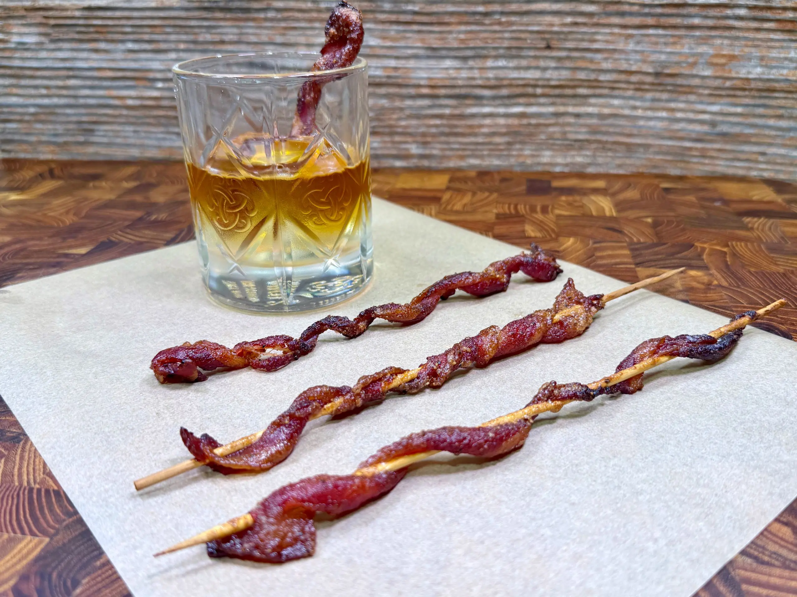 A glass of whiskey with a slice of bacon inside sits on a napkin, next to three skewers of twisted, cooked bacon on a wooden surface.