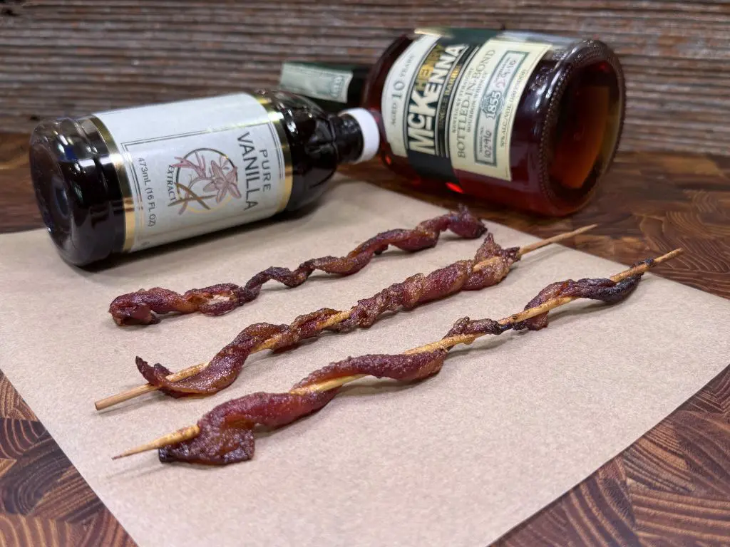 Three twisted strips of candied vanilla bourbon bacon on wooden skewers rest on parchment paper, with bottles of vanilla extract and McKenna bourbon lying on their sides in the background on a wooden surface.