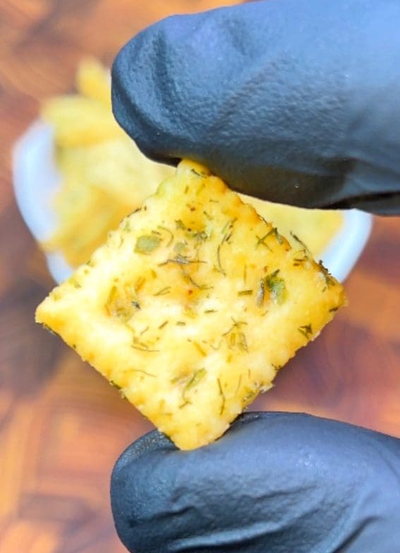 A gloved hand holds a square Saltine cracker topped with fill-picle flavored herbs and butter above a blurred background, which includes a bowl of similar crackers on a wooden surface.