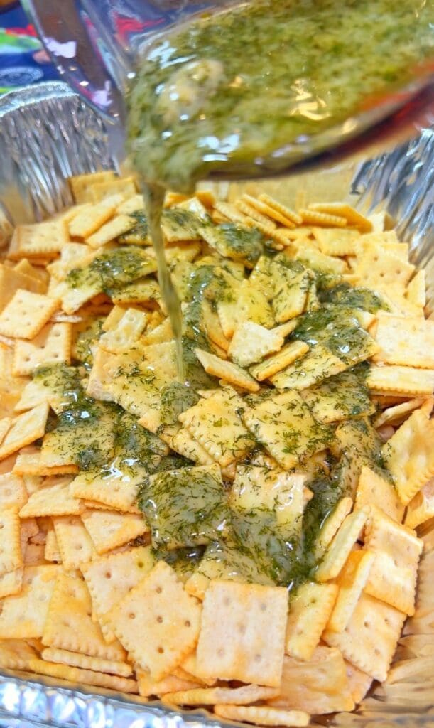 A baking tray filled with square Saltine crackers being drizzled with a green, dill-pickle, dill and butter sauce to make dill pickle crackers.