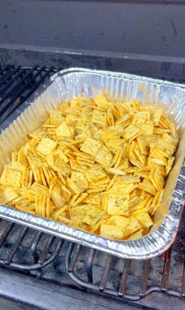 A foil pan filled with seasoned cdill pickle saltines rackers is placed on a grill, with a layer of scattered green herbs visible on top.
